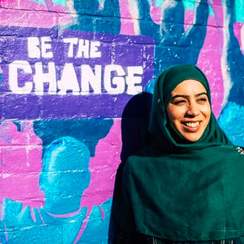 A woman stood in front of a colourful wall with the words 'Be the Change' written on it